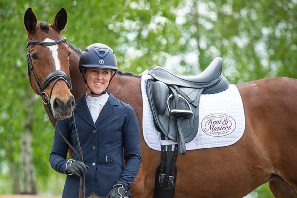 A woman in Kent & Masters Competition Series Monoflap Dressage gear standing next to a brown horse.